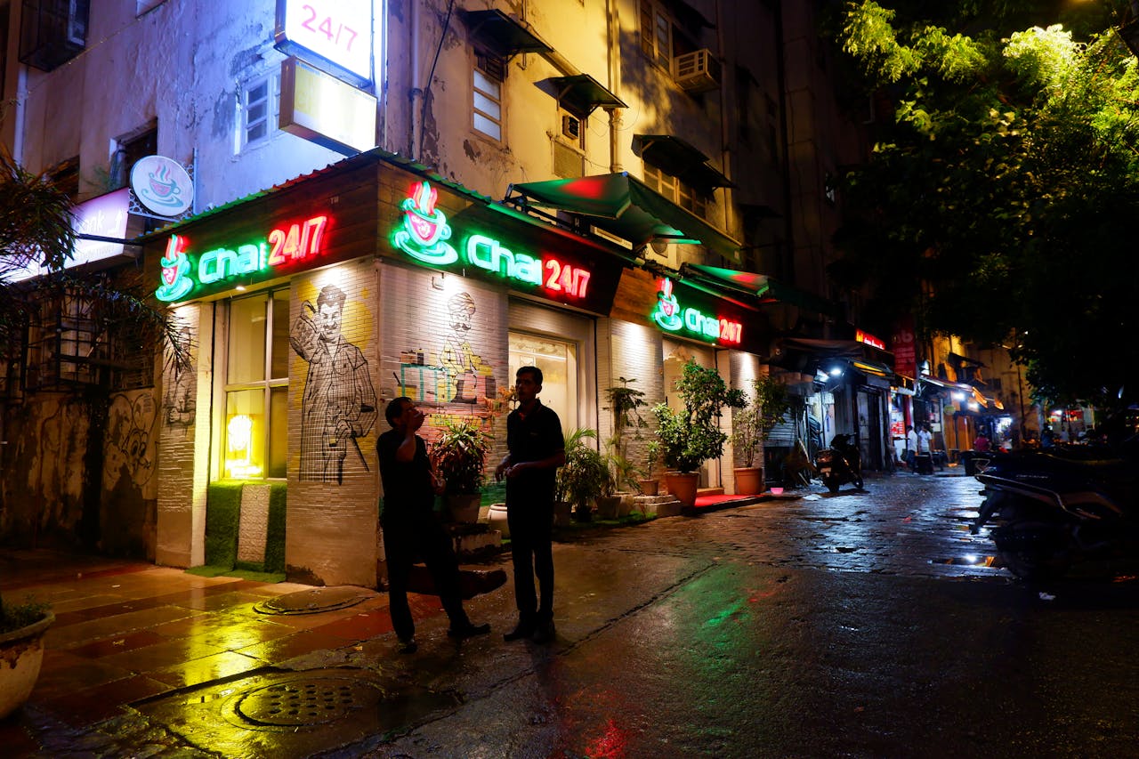 A vibrant street scene at night featuring a glowing cafe storefront and two adults conversing.